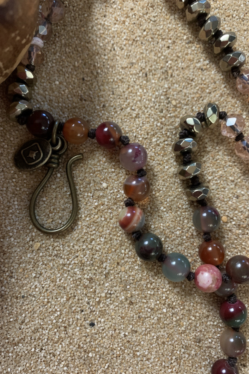 A close-up of a beaded necklace with faceted crystal beads in earthy tones, dark gray agate-like beads, and an antique bronze hook clasp
