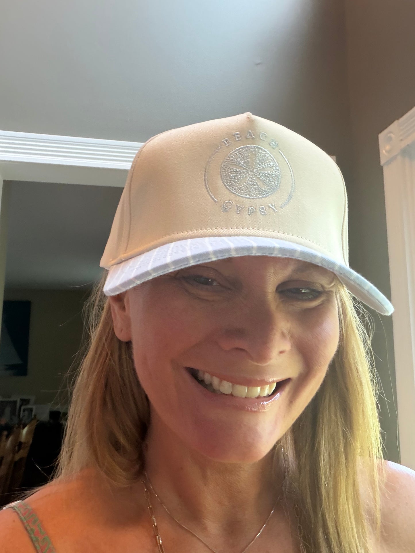 A woman wearing a beige trucker-style cap with a white brim and a Beach Gypsy embroidered logo on the front, photographed indoors.
