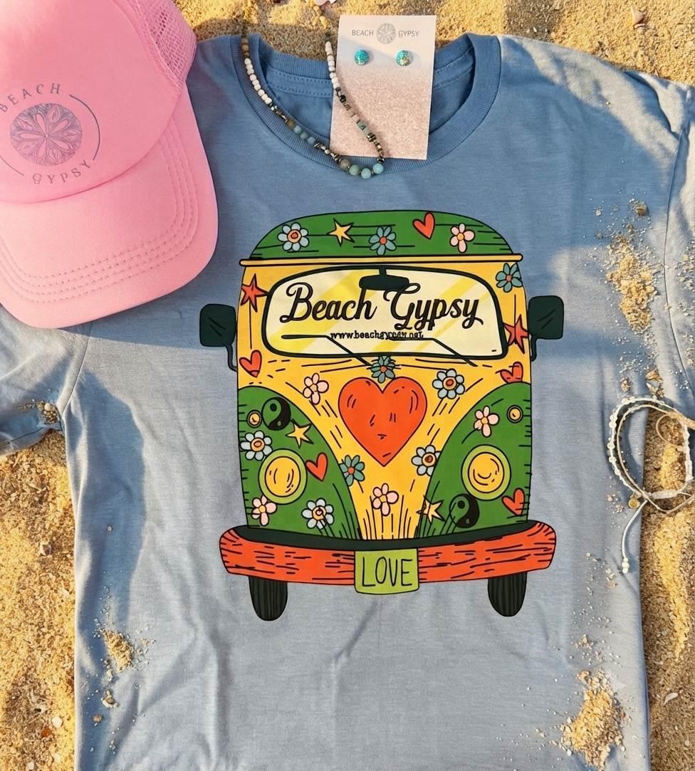 A light blue graphic T-shirt laid flat on sand, featuring a colorful retro van illustration with the words 'Beach Gypsy' and 'LOVE.' A pink mesh cap, beaded necklaces, and bracelets are placed around it as styling props.