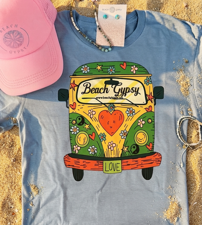 A light blue graphic T-shirt laid flat on sand, featuring a colorful retro van illustration with the words 'Beach Gypsy' and 'LOVE.' A pink mesh cap, beaded necklaces, and bracelets are placed around it as styling props.