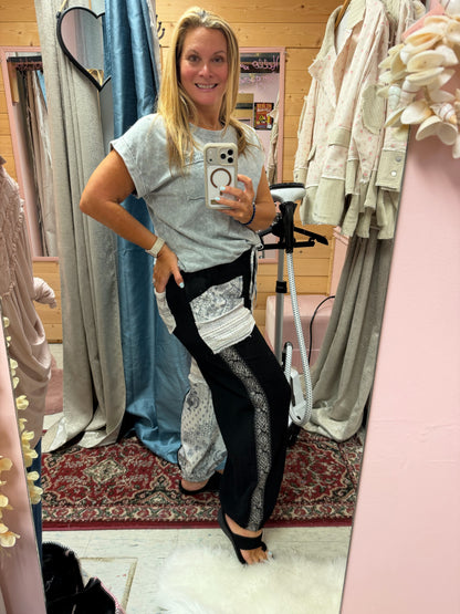 A woman takes a mirror selfie in a boutique showing off a pair of black wide-leg pants with silver patterned side panels and a white textured waistband and elastic pant cuffs, paired with a light gray t-shirt.
