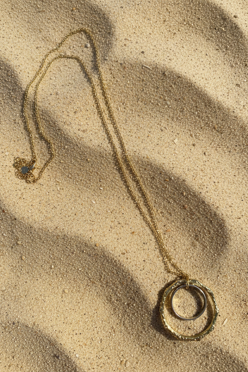 A gold chain necklace with a large circular pendant featuring a textured outer ring and a clear glass insert, displayed on a sand  background.