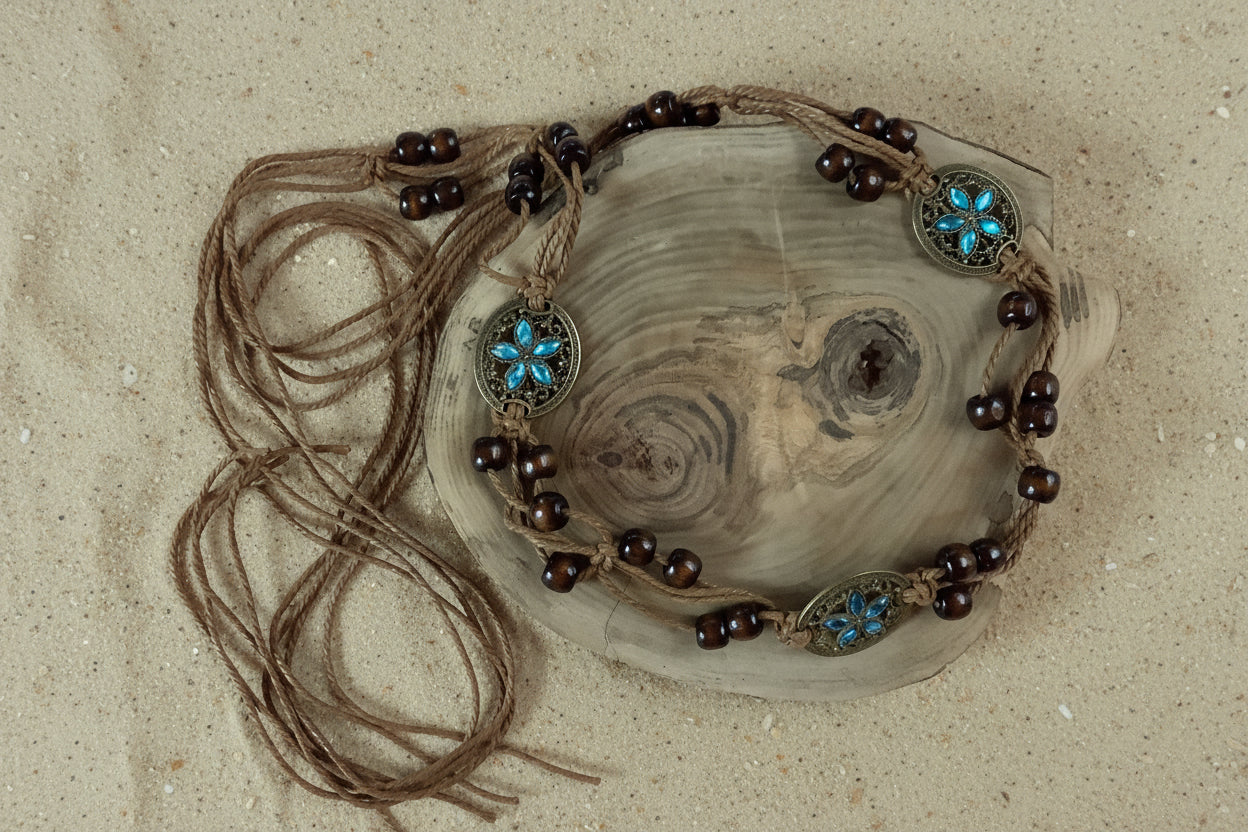A bohemian-style beaded necklace with dark brown wooden beads, brown cord, and turquoise blue starburst metal accents, displayed on a rustic wood slice against a sand background.
