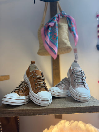 sneakers displayed on a wooden shelf in a boutique setting. The left pair is brown suede with white rubber soles, and the right pair is light pink suede with white rubber soles.