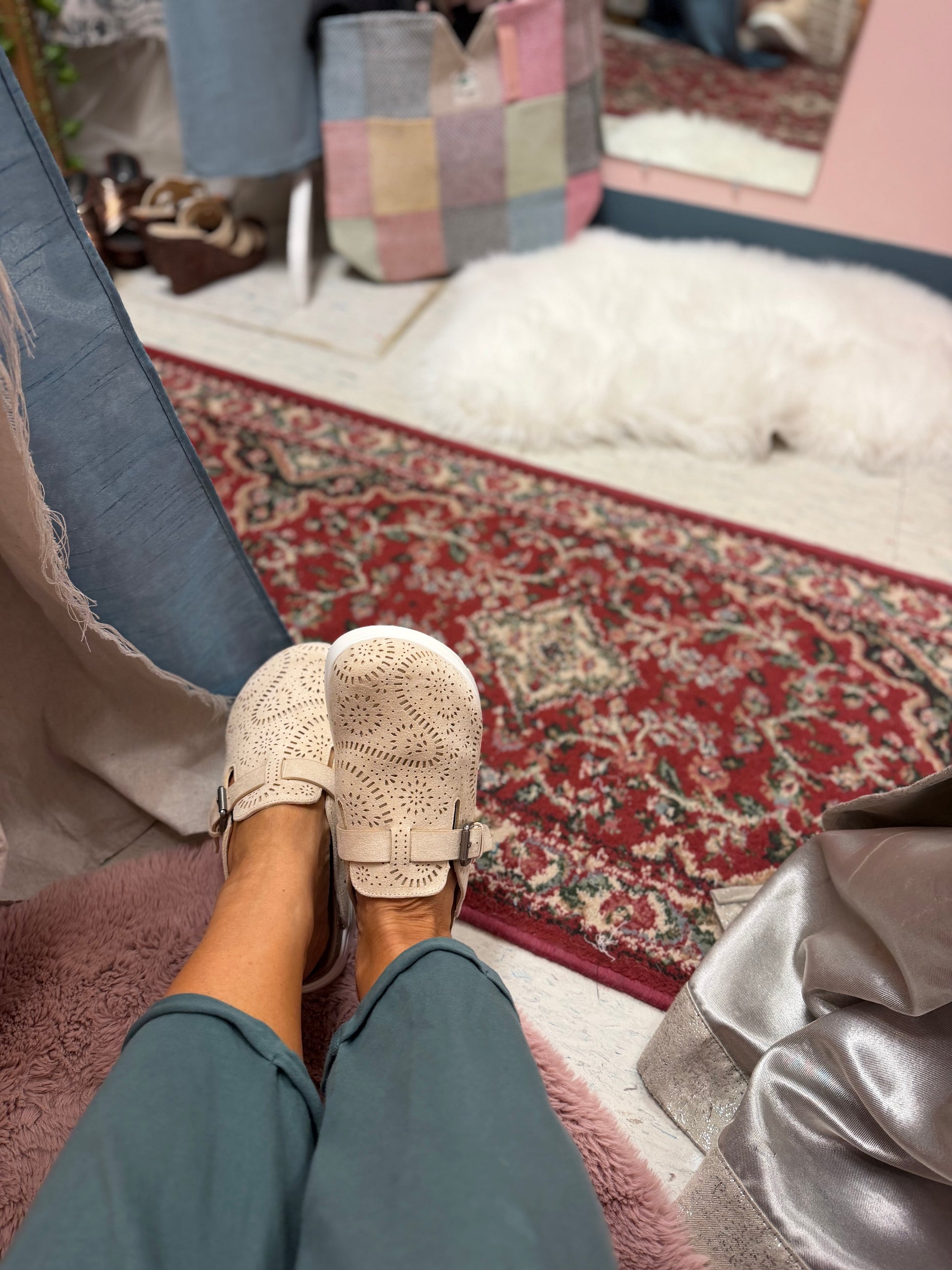 A person wearing cream-colored crochet slip-on shoes with buckle straps and teal pants, sitting on a pink plush chair in a room with a red patterned rug and a white shag rug.