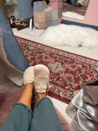 A person wearing cream-colored crochet slip-on shoes with buckle straps and teal pants, sitting on a pink plush chair in a room with a red patterned rug and a white shag rug.
