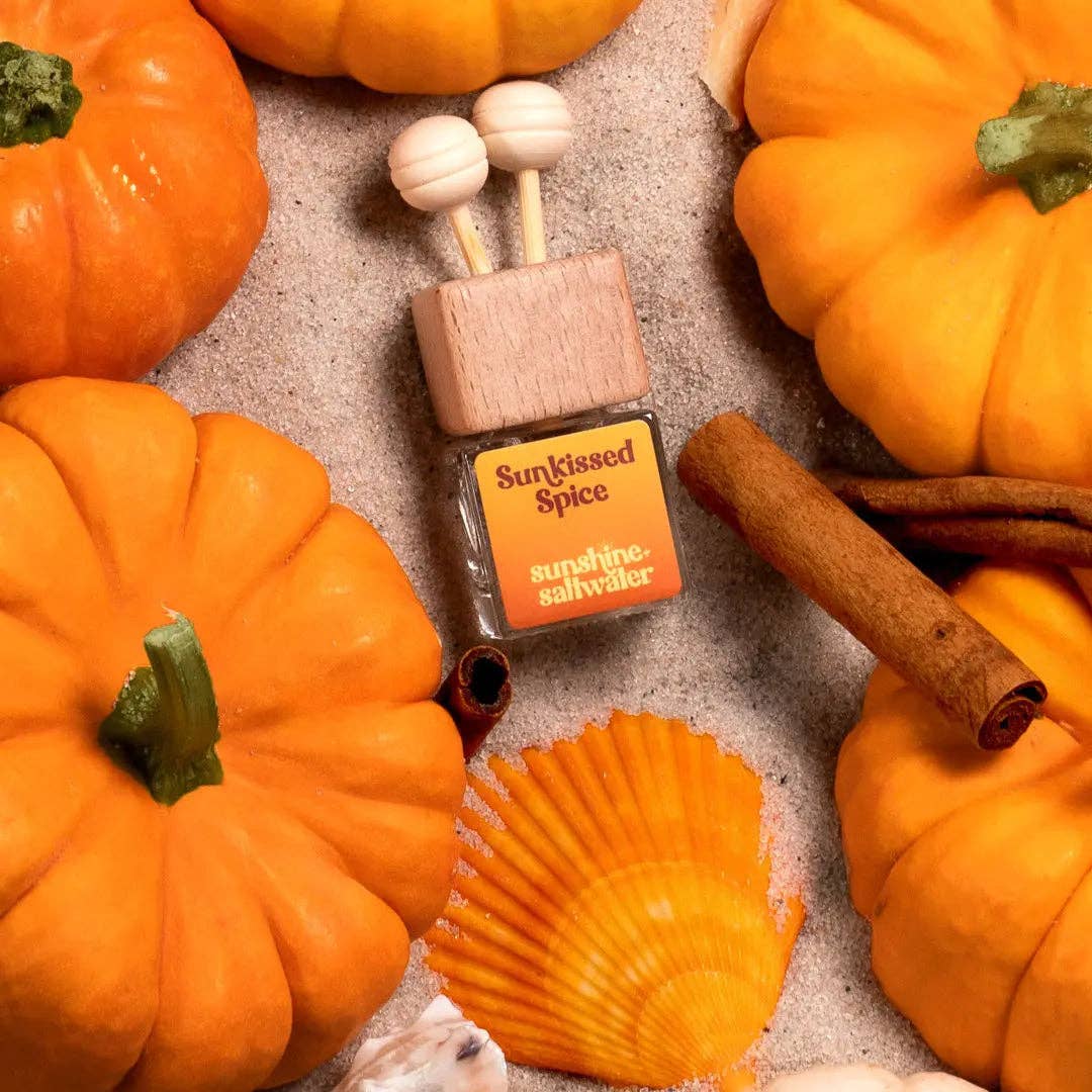 A bottle of Sunshine Saltswayer 'Sunkissed Spice' diffuser oil surrounded by decorative autumn elements including orange pumpkins, cinnamon sticks, a seashell, and garlic on a textured neutral background.