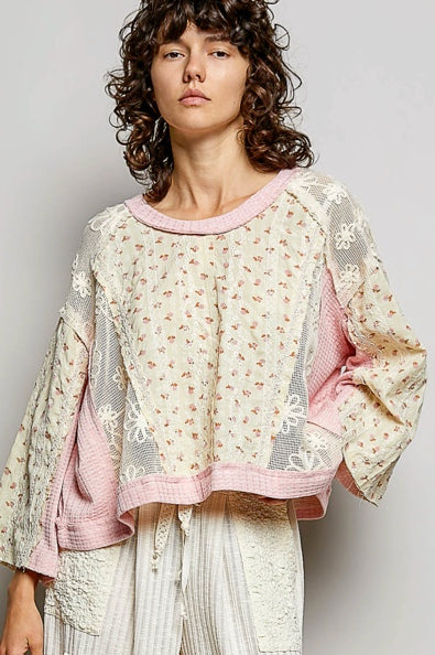 A model wearing a cropped, patchwork-style long-sleeve top with a cream floral print, pink trim, and sheer mesh sleeves, paired with cream pleated trousers, against a plain light gray background.