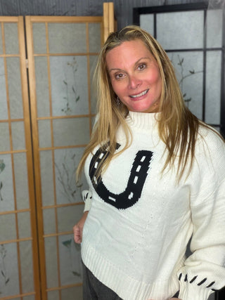 A woman with blonde hair wearing a cream-colored knit sweater with a black embroidered horseshoe design on the front, standing in front of a wooden folding screen.