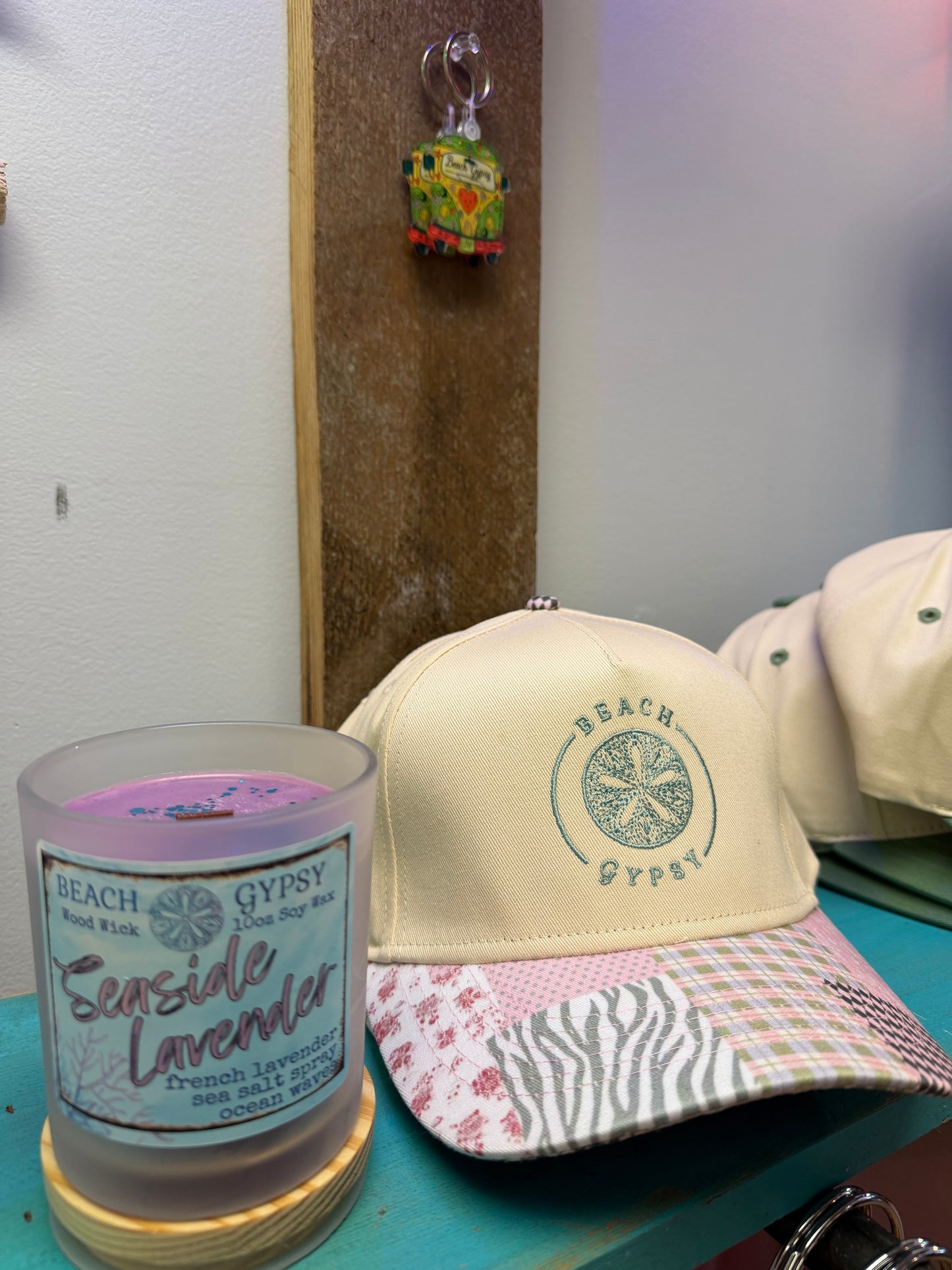 A cream-colored Beach Gypsy trucker cap with a multicolor patterned visor sits on a teal table next to a 'Seaside Lavender' scented candle. The cap features a green circular logo on the front panel.