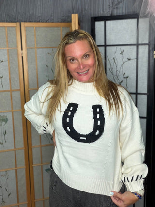 A woman with blonde hair wearing a cream-colored knit sweater with a black horseshoe graphic on the chest, standing in front of a decorative folding screen.