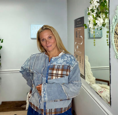 A woman wearing a light blue distressed patchwork zip-up jacket with denim, floral, and plaid panels, standing in an indoor setting with a mirror and wall decor in the background.