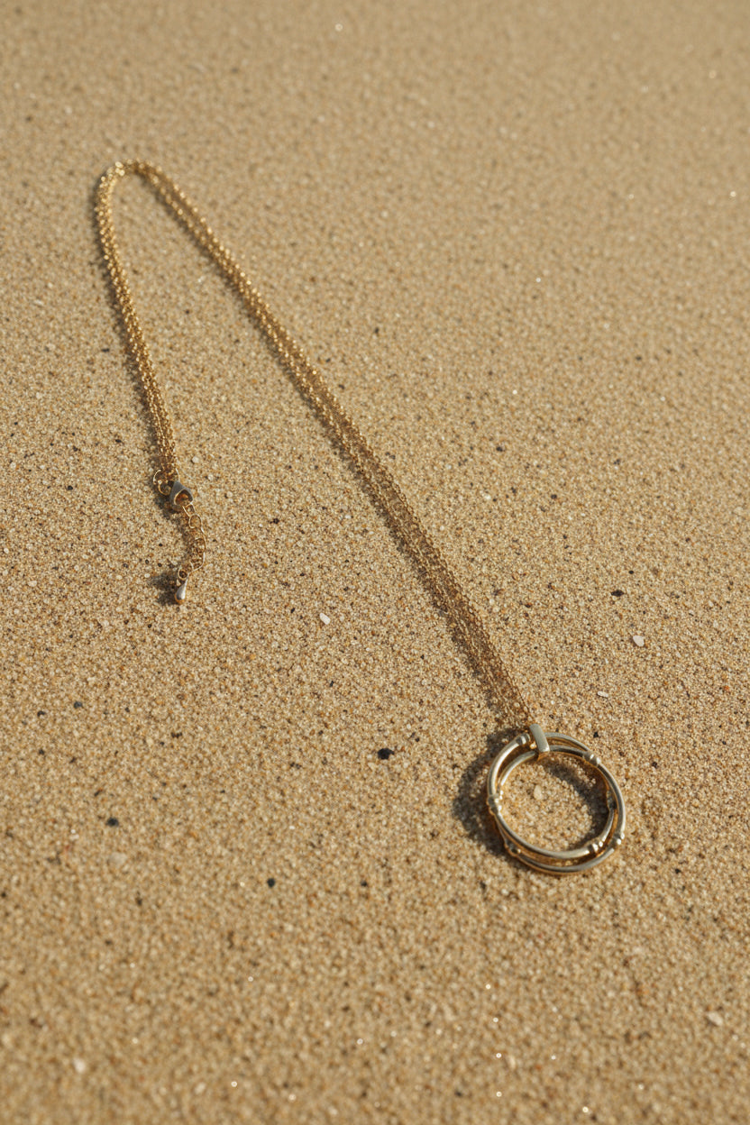 A gold-toned chain necklace with a large circular ring pendant and an adjustable sliding clasp, displayed on a sand  background.