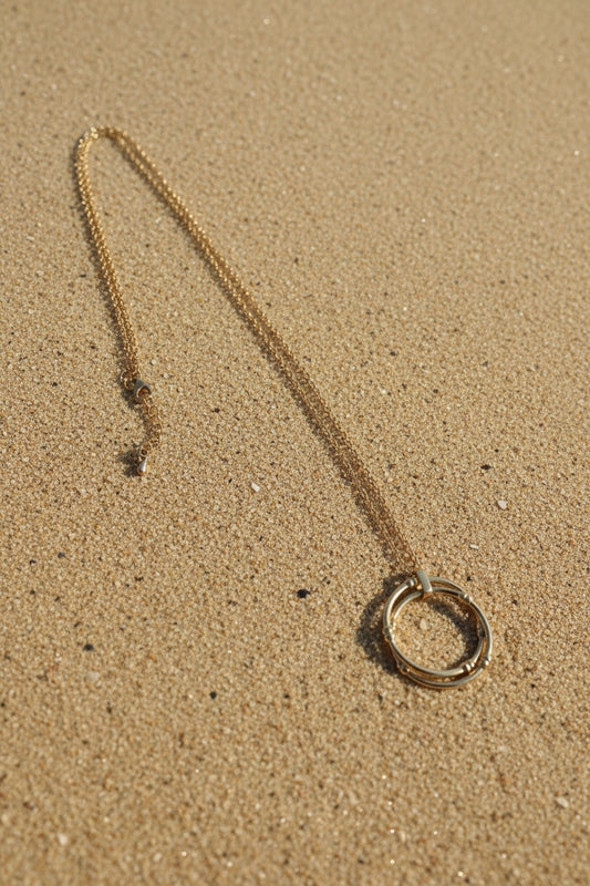 A gold-toned chain necklace with a large circular ring pendant and an adjustable sliding clasp, displayed on a sand  background.