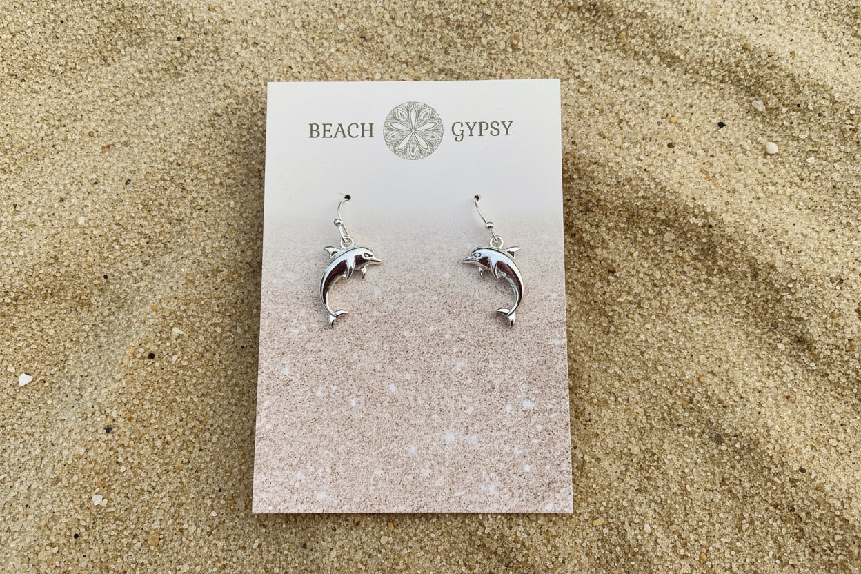 Dolphin-shaped earrings on a 'Beach Gypsy' branded card against a sand background