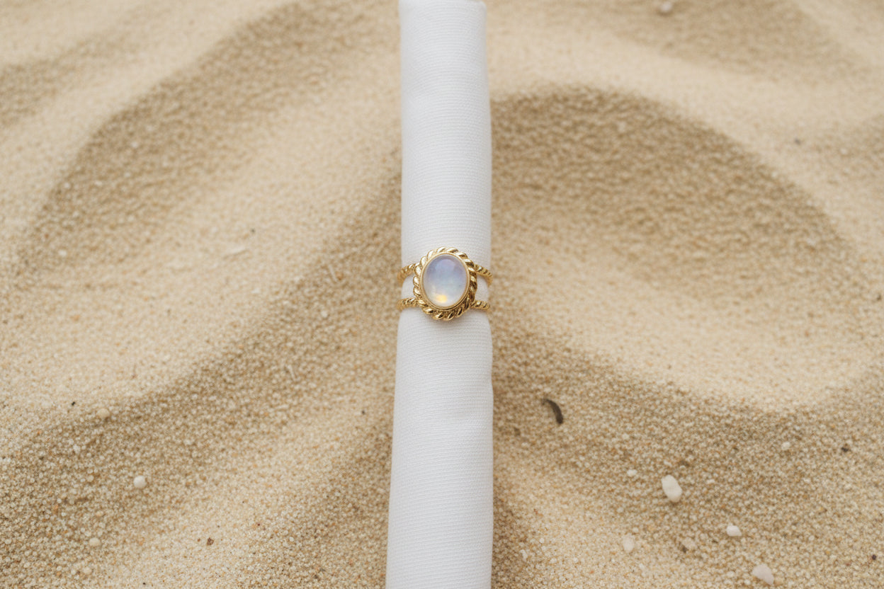 A gold ring with a round, pink stone, displayed on a white fabric ring holder over a brown woven cushion.