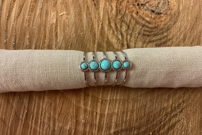A multi-band silver ring set with five turquoise cabochon stones of varying sizes, displayed on a rolled cream fabric sleeve over a rustic wooden surface.