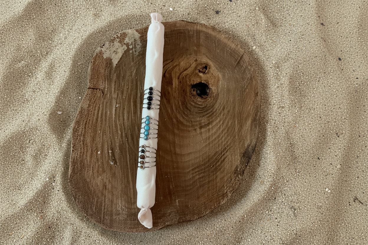 A white wrapped candle with colorful beads and silver wire accents, displayed vertically on a rustic round wooden slice against a sand background.