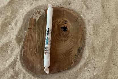 A white wrapped candle with colorful beads and silver wire accents, displayed vertically on a rustic round wooden slice against a sand background.
