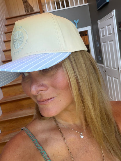 A woman wearing a light beige baseball cap with a light blue and white striped brim, photographed indoors in front of a staircase and doors.