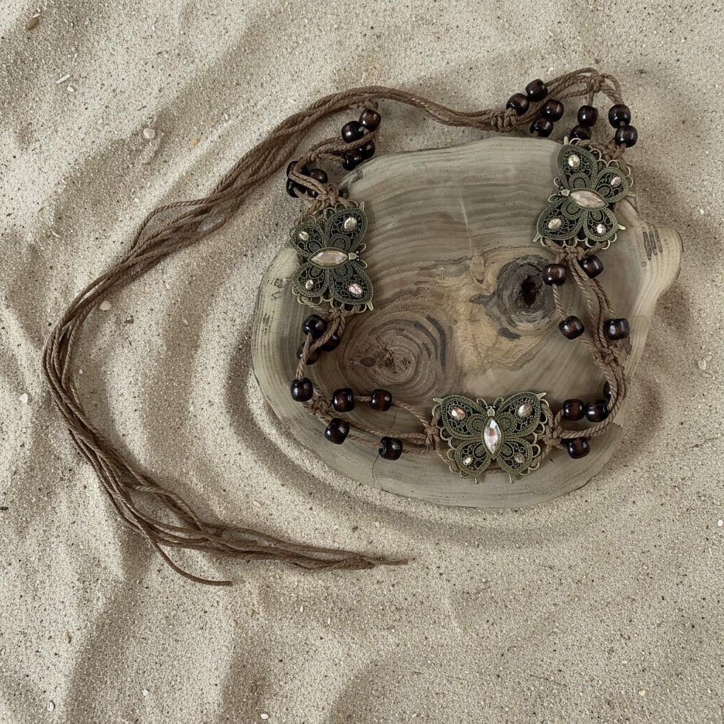 A decorative macrame shoulder bag with dark brown beads and bronze butterfly embellishments, displayed on a rustic wood slice prop against a sand  background.