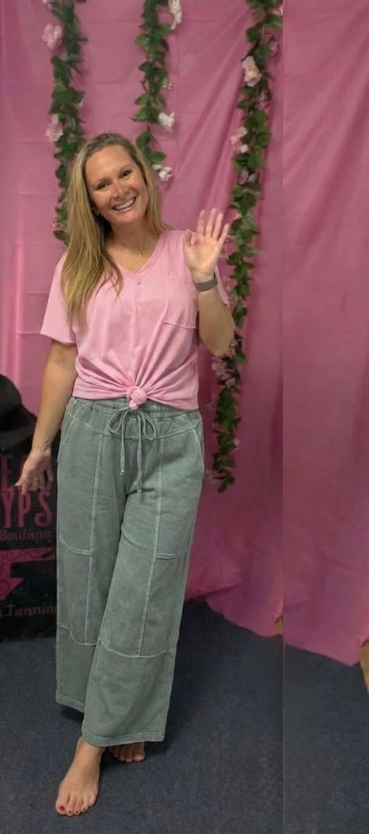 Woman in pink top and green pants waving in front of a pink curtain with floral decorations.