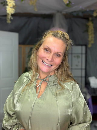 A smiling blonde woman wearing a shiny sage green satin blouse with a tie-neck detail, photographed indoors in what appears to be a decorated event space or tent.