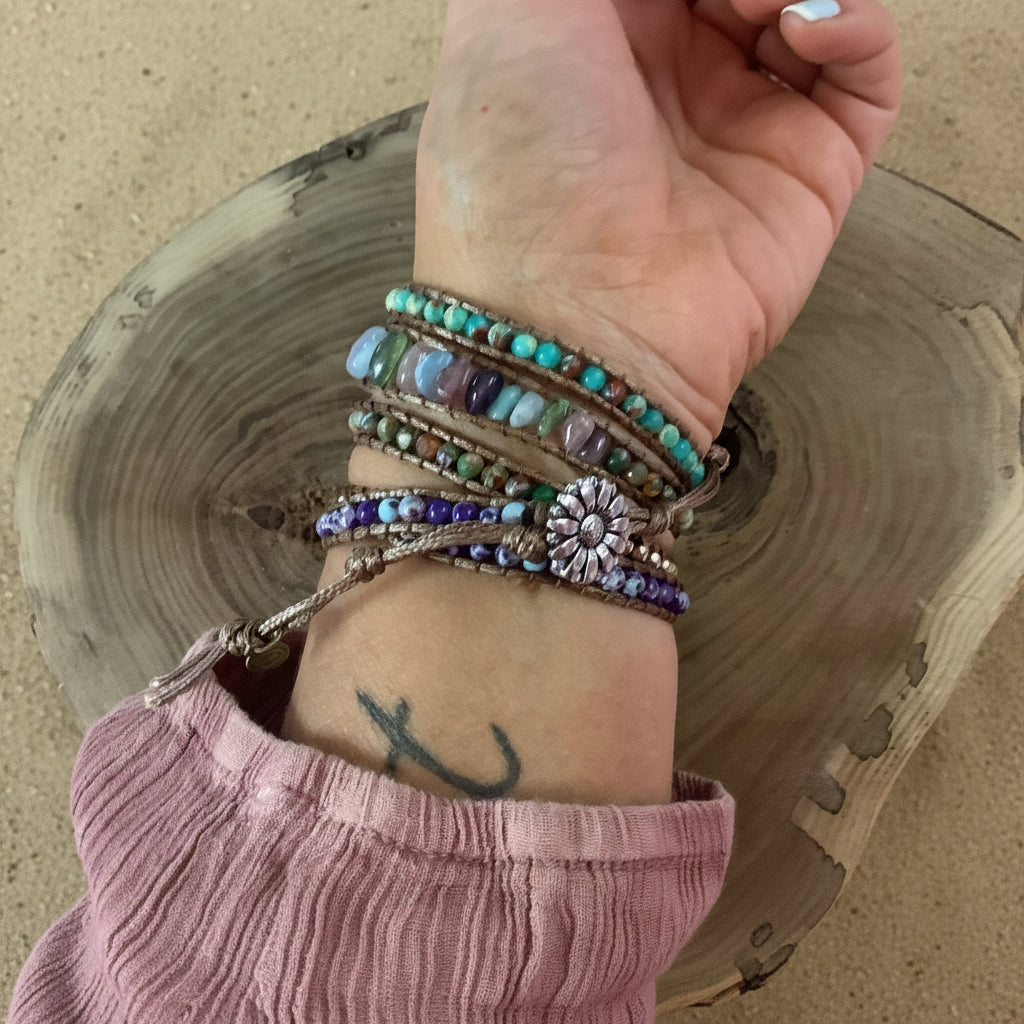 A close-up of a person's wrist wearing multiple beaded bracelets, including turquoise, purple, green, and blue beads with a silver-toned flower charm, resting on a rustic wood slice against a sand background.