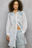 Woman wearing a white floral embroidered shirt and striped pants against a gray background