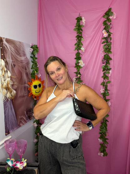 A woman posing in a white halter top and gray pants, holding a black purse, set against a pink backdrop decorated with floral garlands.
