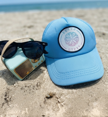 Baby blue trucker hat with Beach Gypsy logo on a sandy surface next to a blue radio and a pair of sunglasses. All can be bought at Beach Gypsy in Berlin, MD!