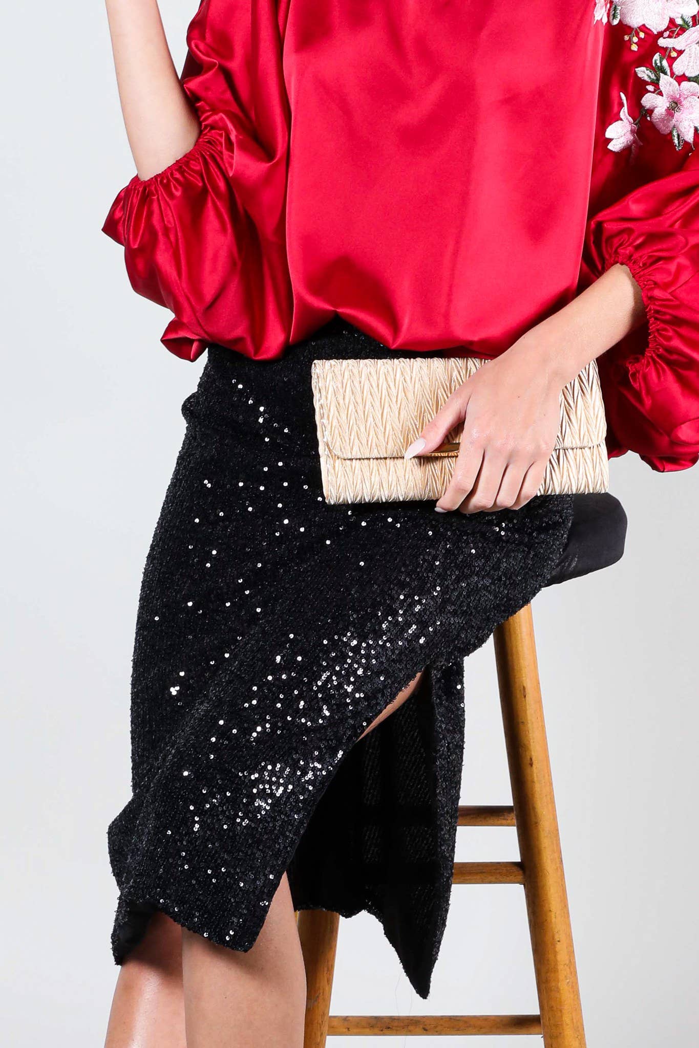 Person wearing a red top and black sequin skirt holding a beige clutch on a white background