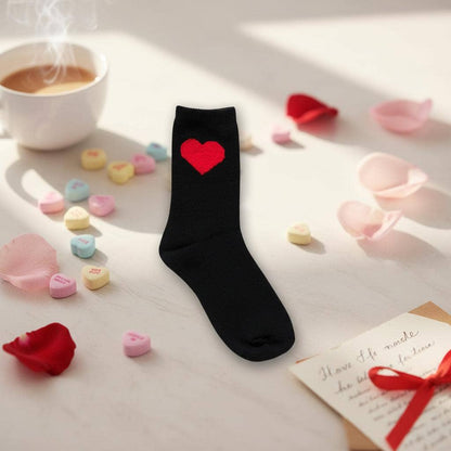 Black sock with a red heart on a surface with coffee, candy hearts, and rose petals.