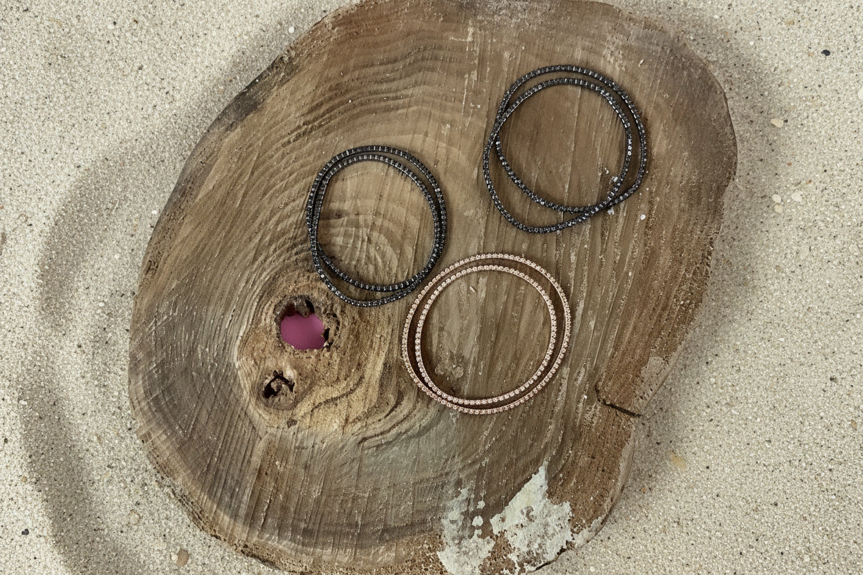 Three beaded bangles in dark gray, black, and bronze tones arranged on a rustic wooden slice against a sand background.