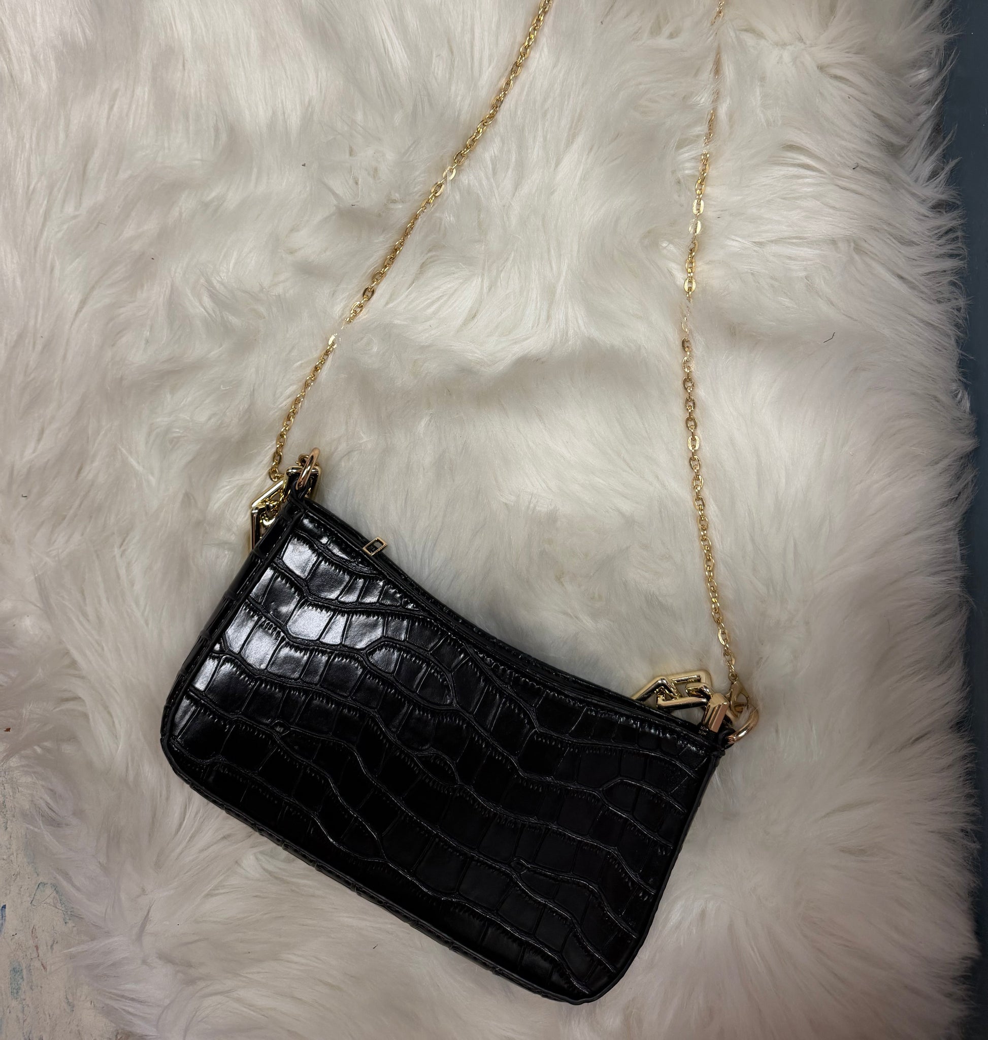 A black crocodile-embossed leather clutch bag with a gold chain strap, resting on a white faux fur surface.