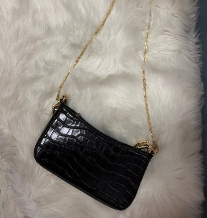 A black crocodile-embossed leather clutch bag with a gold chain strap, resting on a white faux fur surface.