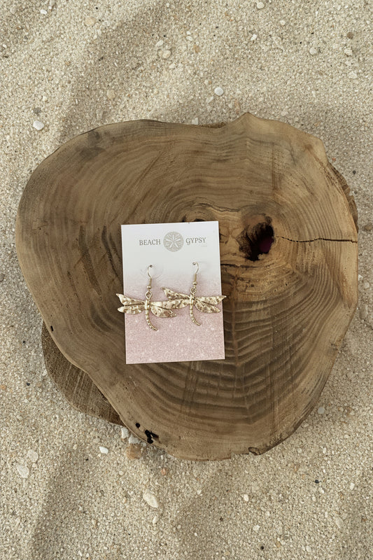 A pair of gold-toned dangle earrings with clear rhinestone details, displayed on a white card labeled 'BEACH GYPSY' and placed on a round natural wood slice against a bright pink background.