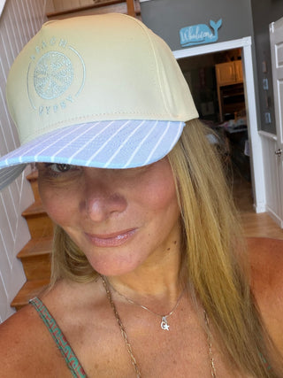 A woman wearing a cream-colored trucker hat with a light blue and white striped brim. The hat features a circular embroidered logo on the front with the text 'beach Gypsy’