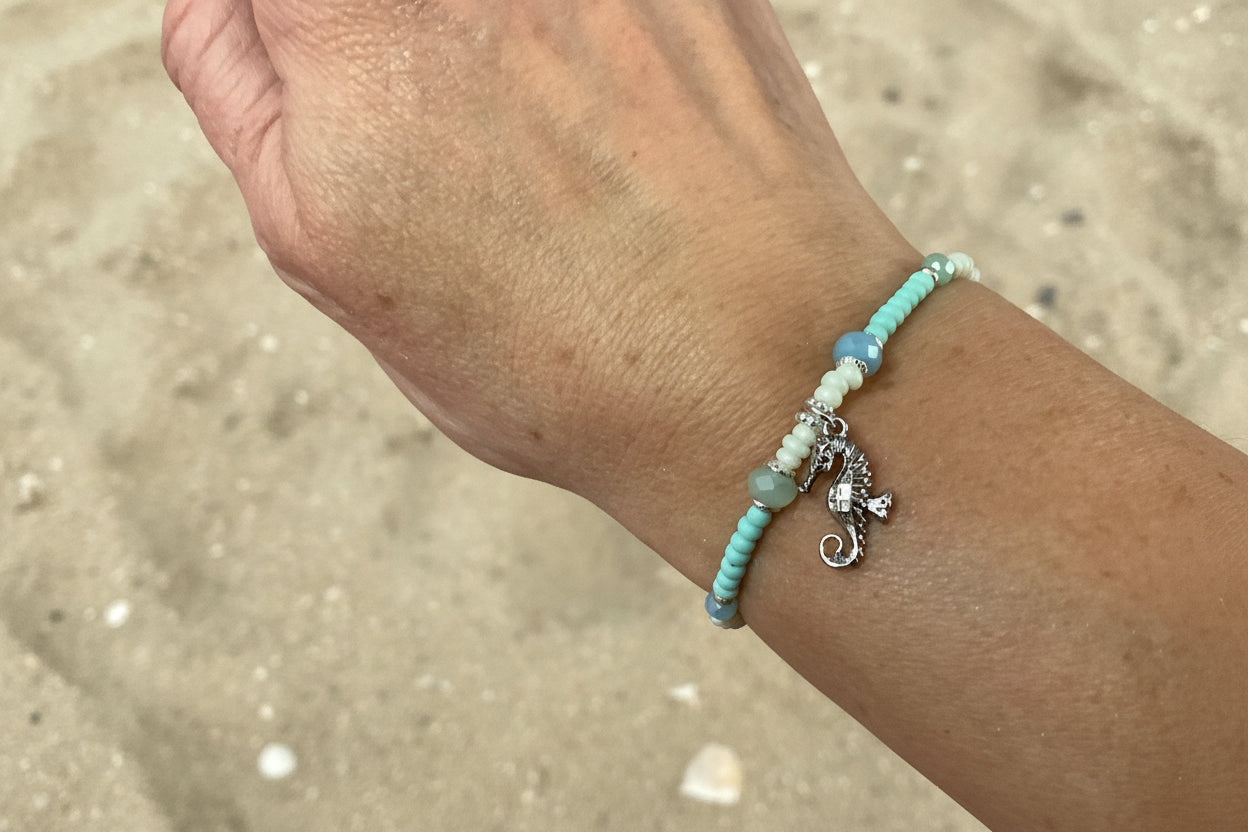 Close-up of a wrist wearing a beaded bracelet with seahorse charm against a sand background
