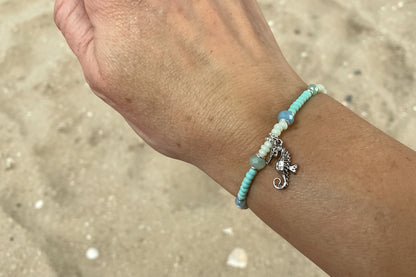 Close-up of a wrist wearing a beaded bracelet with seahorse charm against a sand background