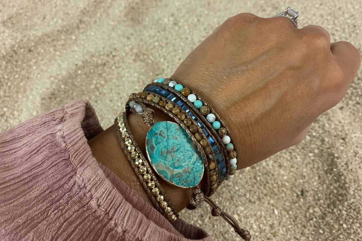 A close-up of a person's wrist wearing a stack of bohemian-style bracelets, featuring a large oval turquoise stone, blue beads, brown leather cords, and gold-toned metal details, set against a sand background.