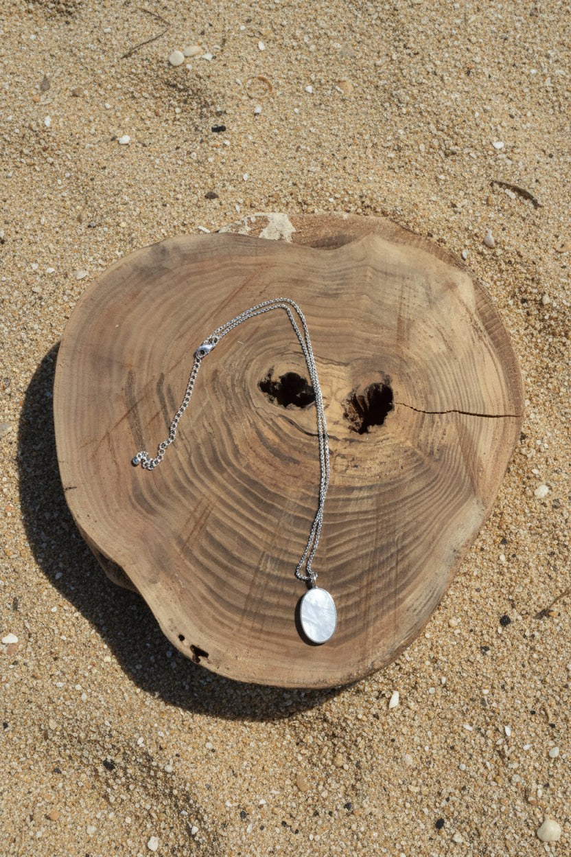A silver chain necklace with an oval white stone pendant, displayed on a rustic wood slice against a sand background.