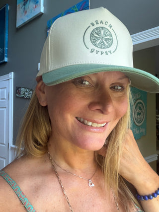 A woman wearing a cream-colored Beach Gypsy trucker hat with a sage green trucker hat and matching green embroidered logo, shown in a close-up lifestyle shot.