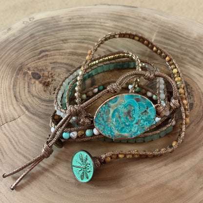 A bohemian multi-strand wrap bracelet featuring turquoise-colored beads, brown cord, a large oval turquoise stone centerpiece, and a small dragonfly charm, displayed on a rustic wood slice background.