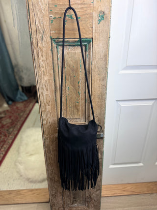 Black Sueded Fringe Crossbody Handbag with long strap and boho fringe, hanging on a rustic wood door – Beach Gypsy