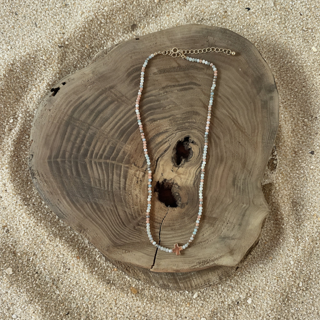 A delicate beaded necklace with small light blue, white, and peach beads draped over a rustic wood slice, set against a sand background.