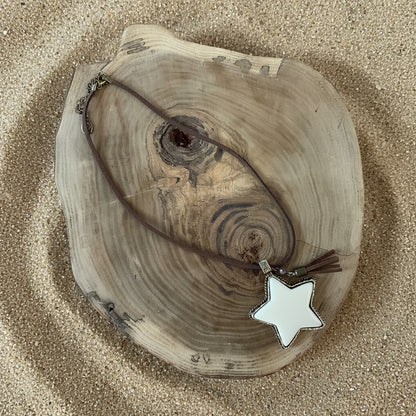 A brown leather cord necklace with a white star-shaped pendant and silver-toned metal accents, displayed on a rustic wood slice against a sand background.