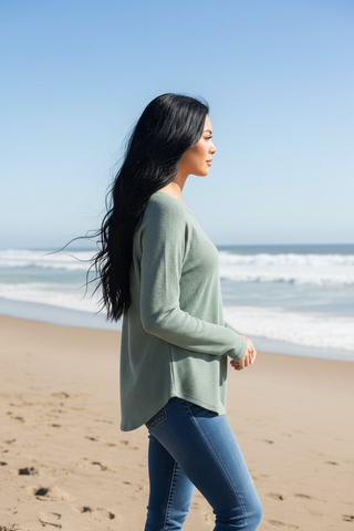 A woman with long black hair walks along a sandy beach wearing a sage green long-sleeve knit top and blue jeans, with the ocean and blue sky in the background.