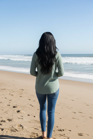 A woman with long black hair stands on a sandy beach facing the ocean, wearing a sage green long-sleeve top and blue jeans.