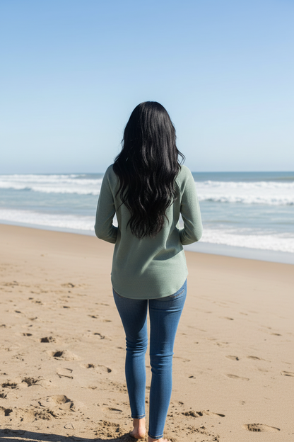 A woman with long black hair stands on a sandy beach facing the ocean, wearing a sage green long-sleeve top and blue jeans.
